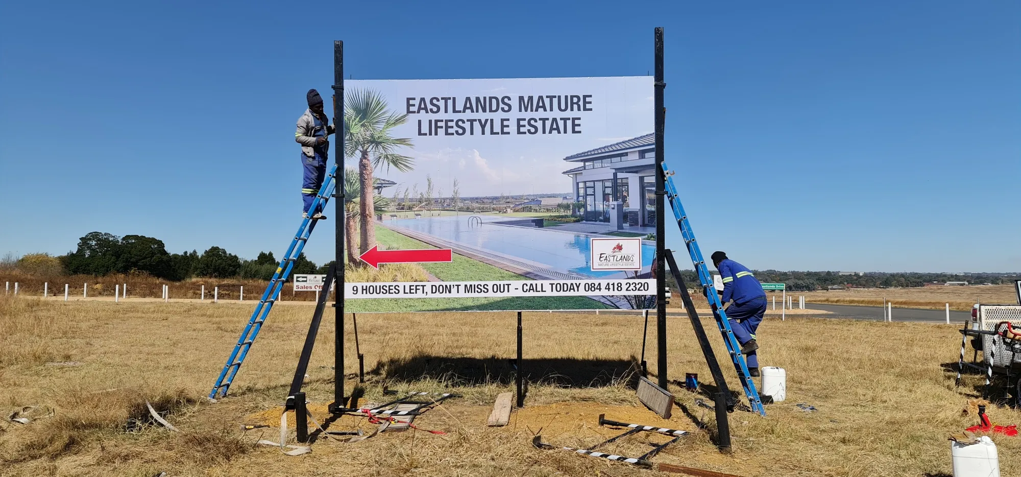 Two installers on ladders fitting a large printed billboard onto steel posts beside a road.