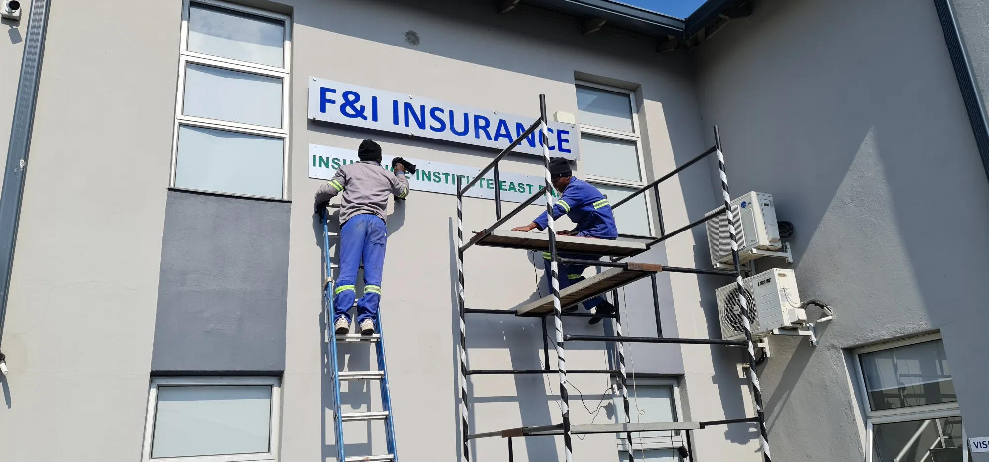 Installers on a ladder and scaffold mounting a lettered sign board onto an office building wall.