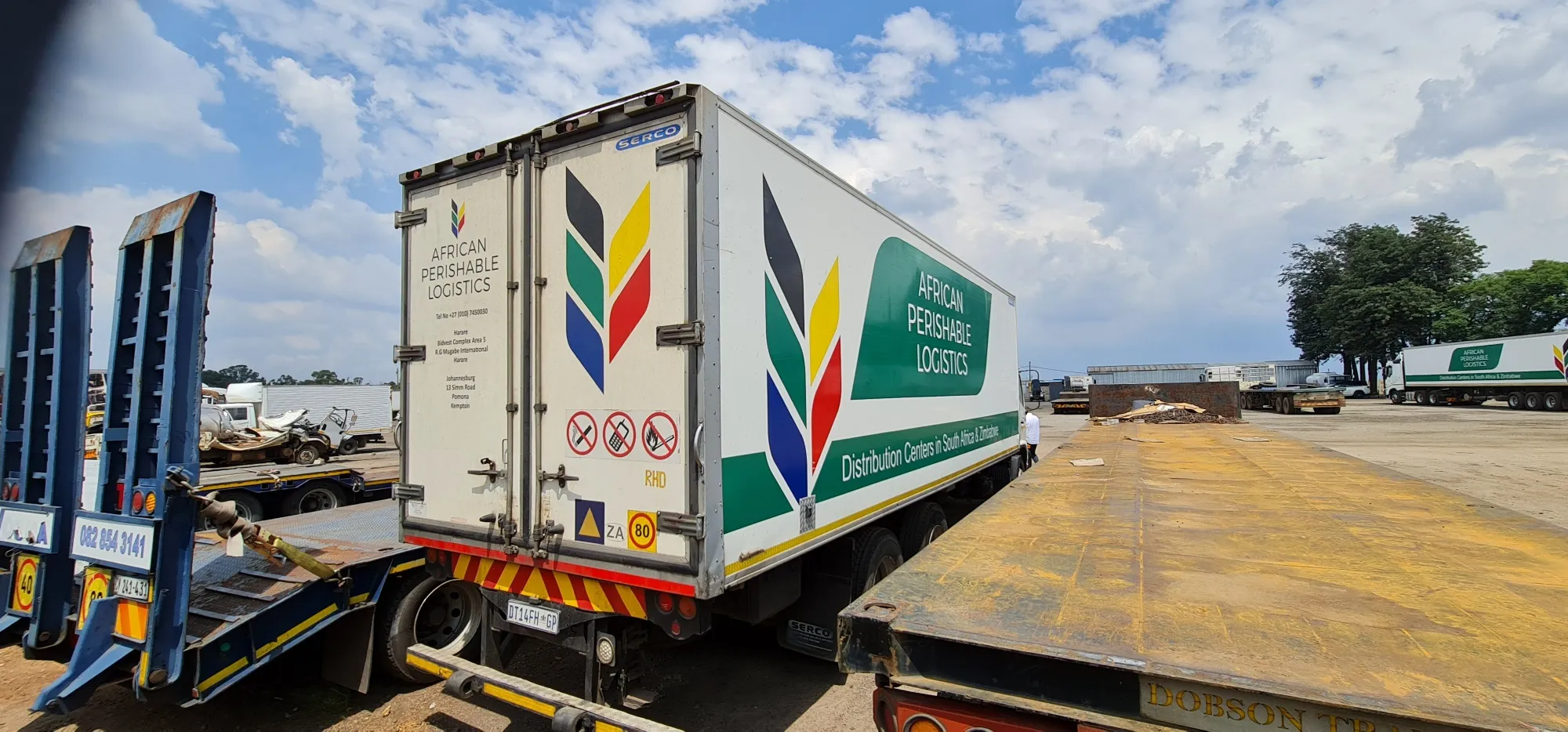 Rear and side view of a branded refrigerated truck in a depot yard, with matching branded trucks behind it.